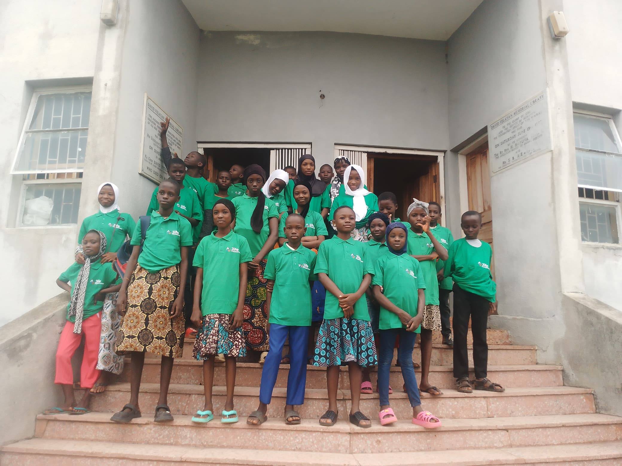 Young people in green JIFE Foundation polo shirts standing together on the steps of the David Folayan Memorial Library.